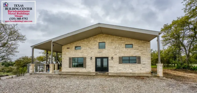 A stone building with a large porch and a gray roof, on a gravel lot under a cloudy sky.
