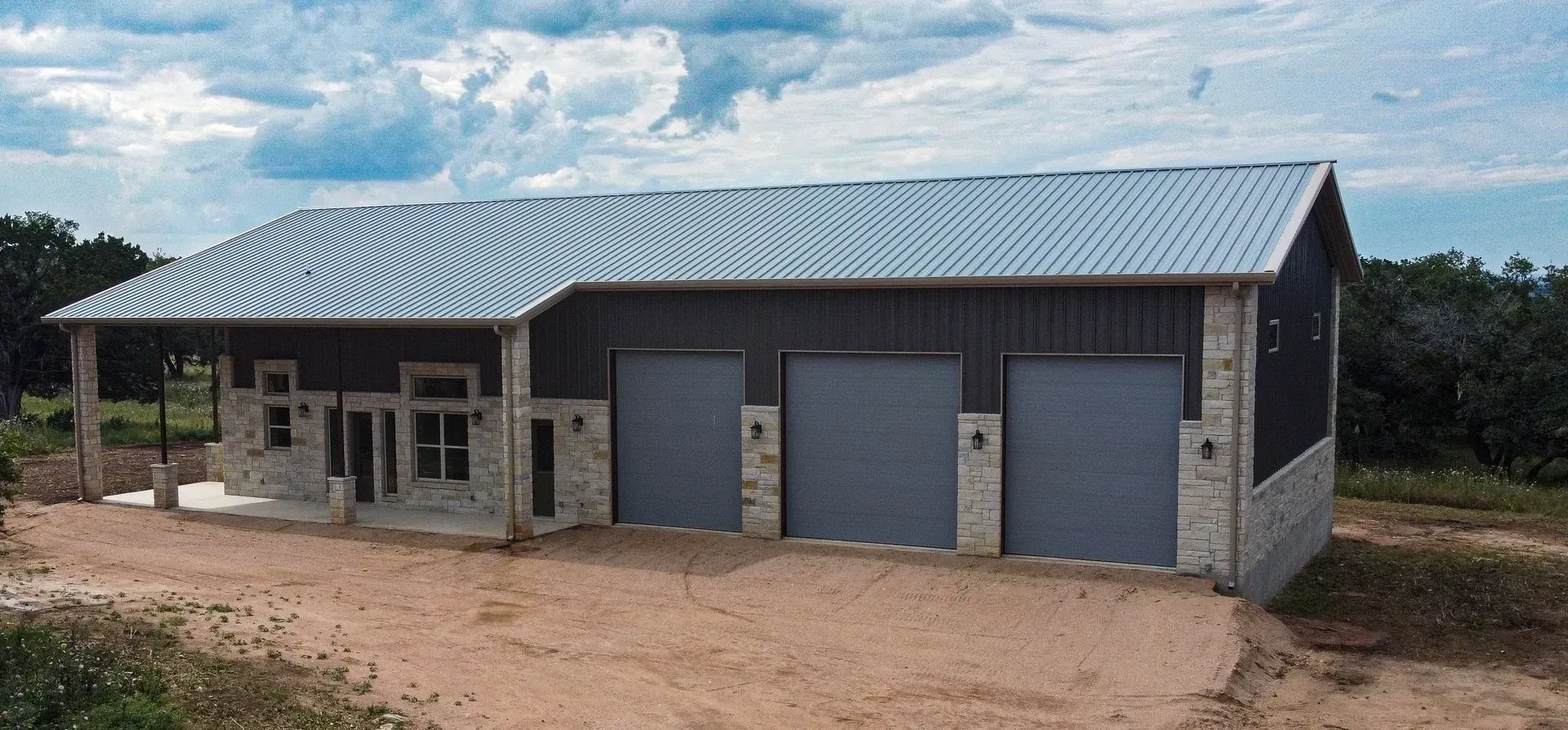 Building with metal roof, three garage doors, stone accents, and a dirt driveway.