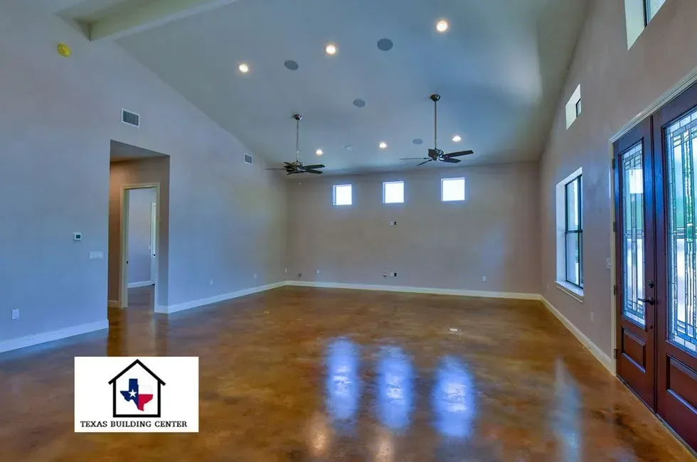 Empty living room with high ceilings, fans, and stained concrete floor; logo in the foreground.