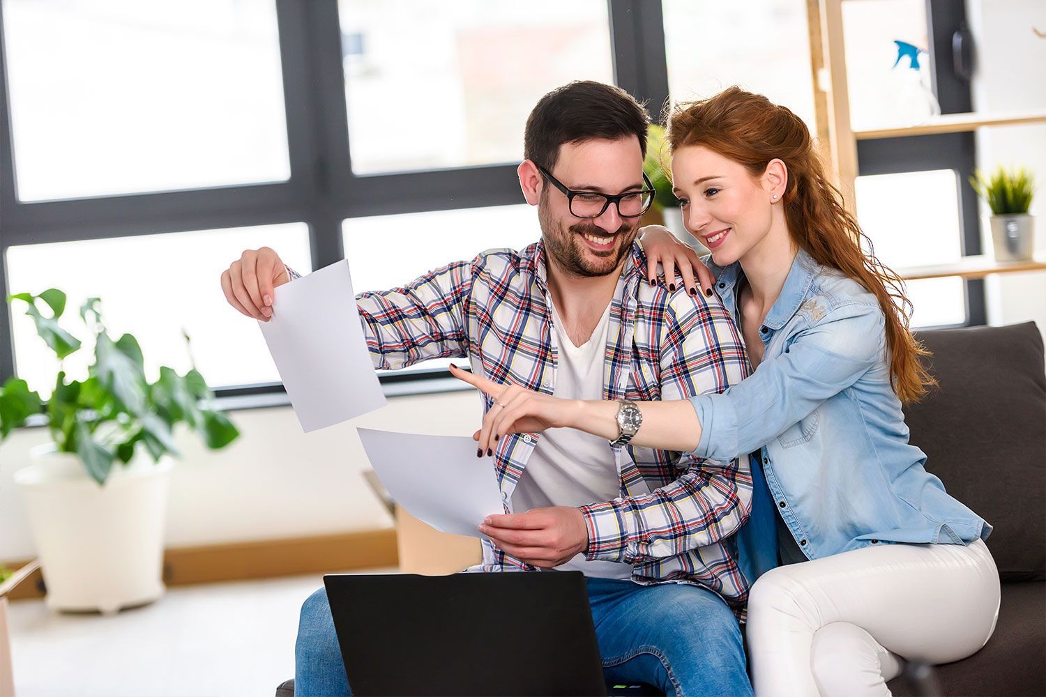 A man and a woman are sitting on a couch looking at papers and a laptop.