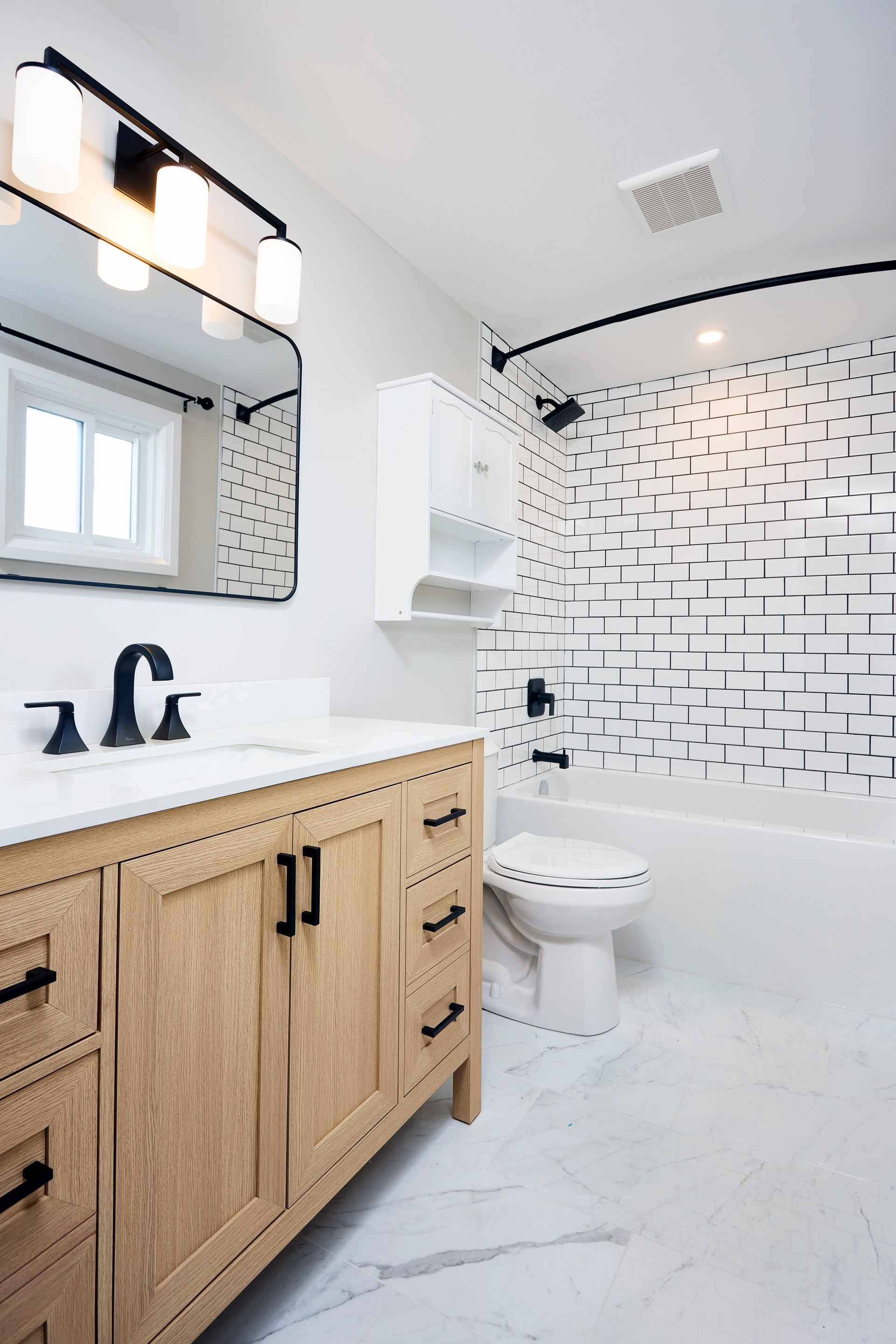 Bathroom with wood vanity, black fixtures, patterned shower wall, white tile floor, and window.