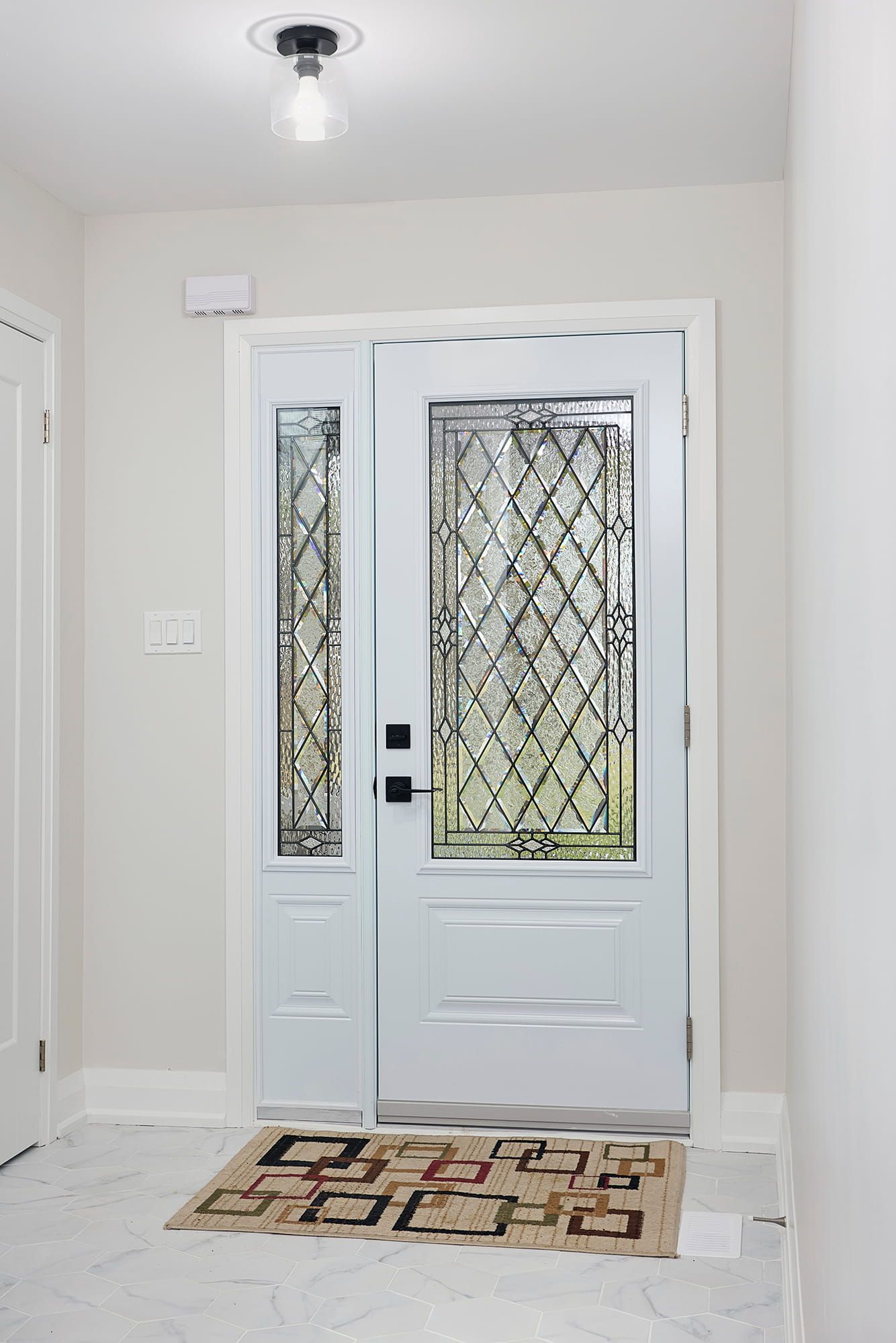 White front door with leaded glass sidelight and doormat, under ceiling light.