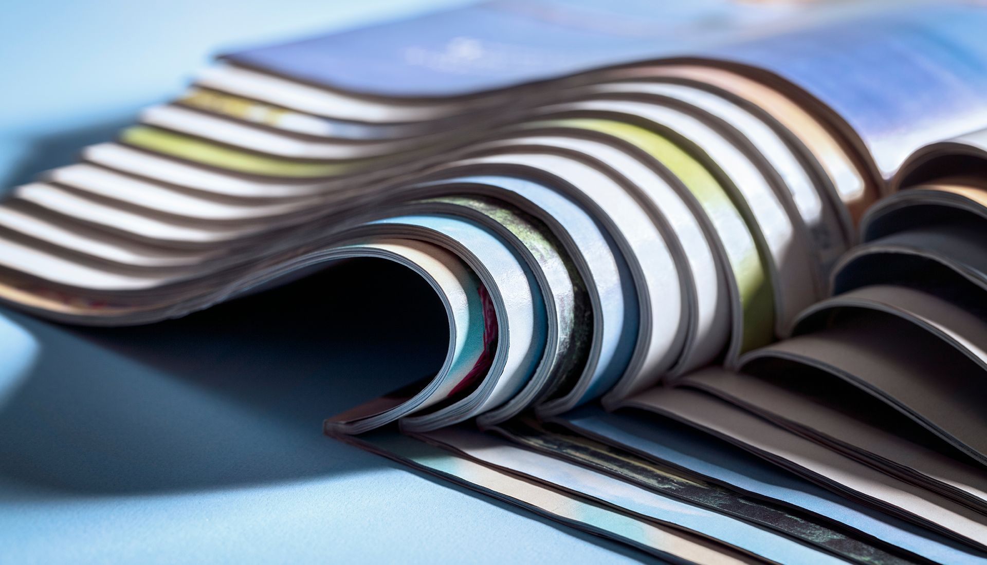 Pile of open magazines, pages displaying varied colors, resting on a light blue surface.
