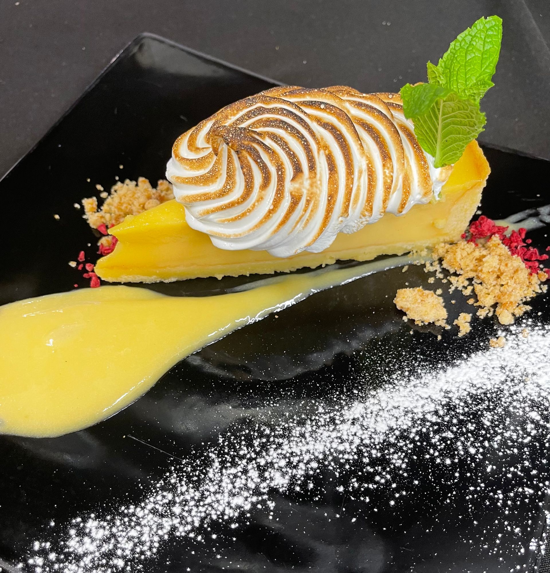 Pavlova dessert with whipped cream, strawberries, and passion fruit sauce on a black plate, garnished with mint.