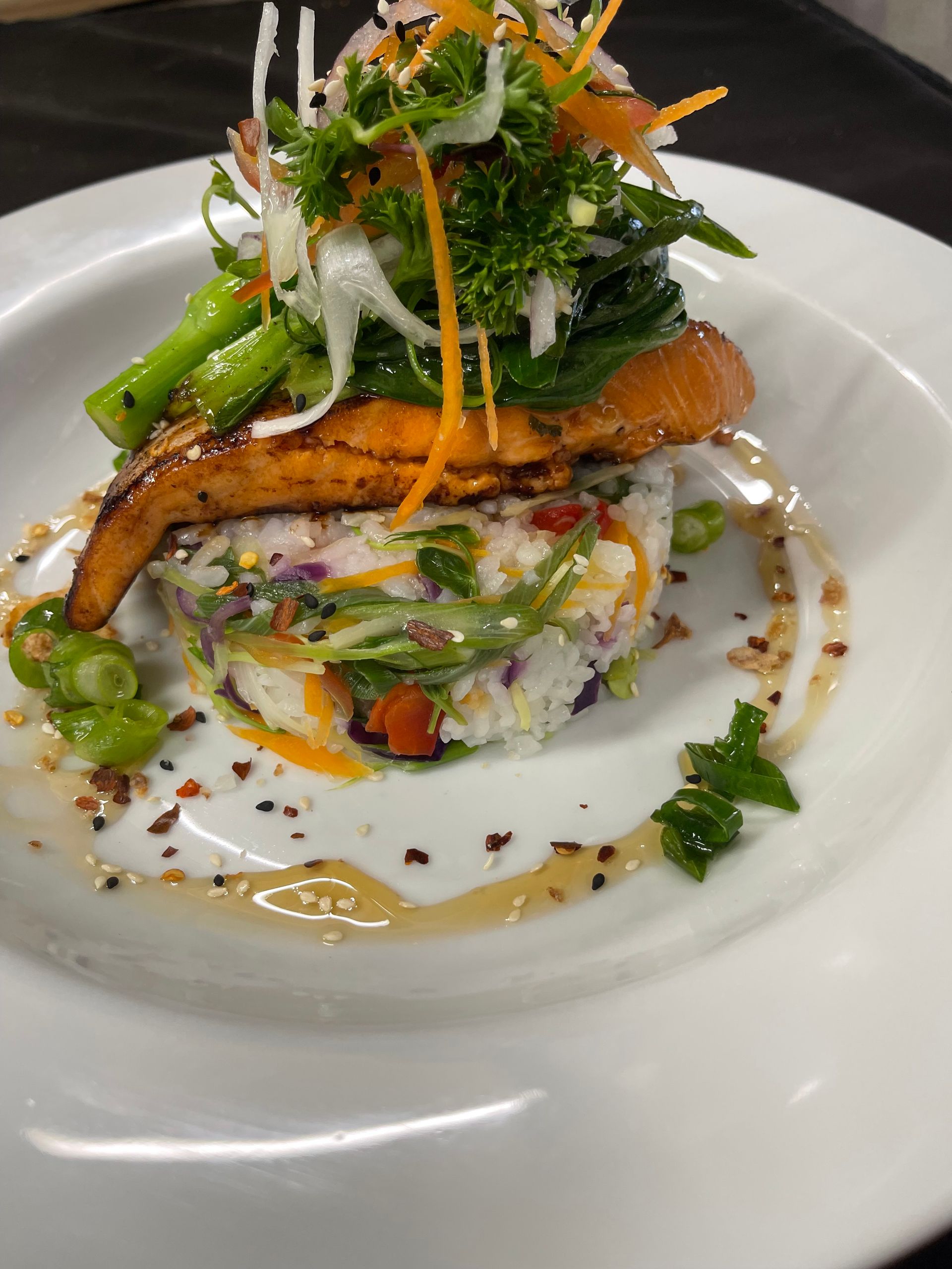 Grilled salmon atop rice and vegetable tower, garnished with greens, on a white plate.