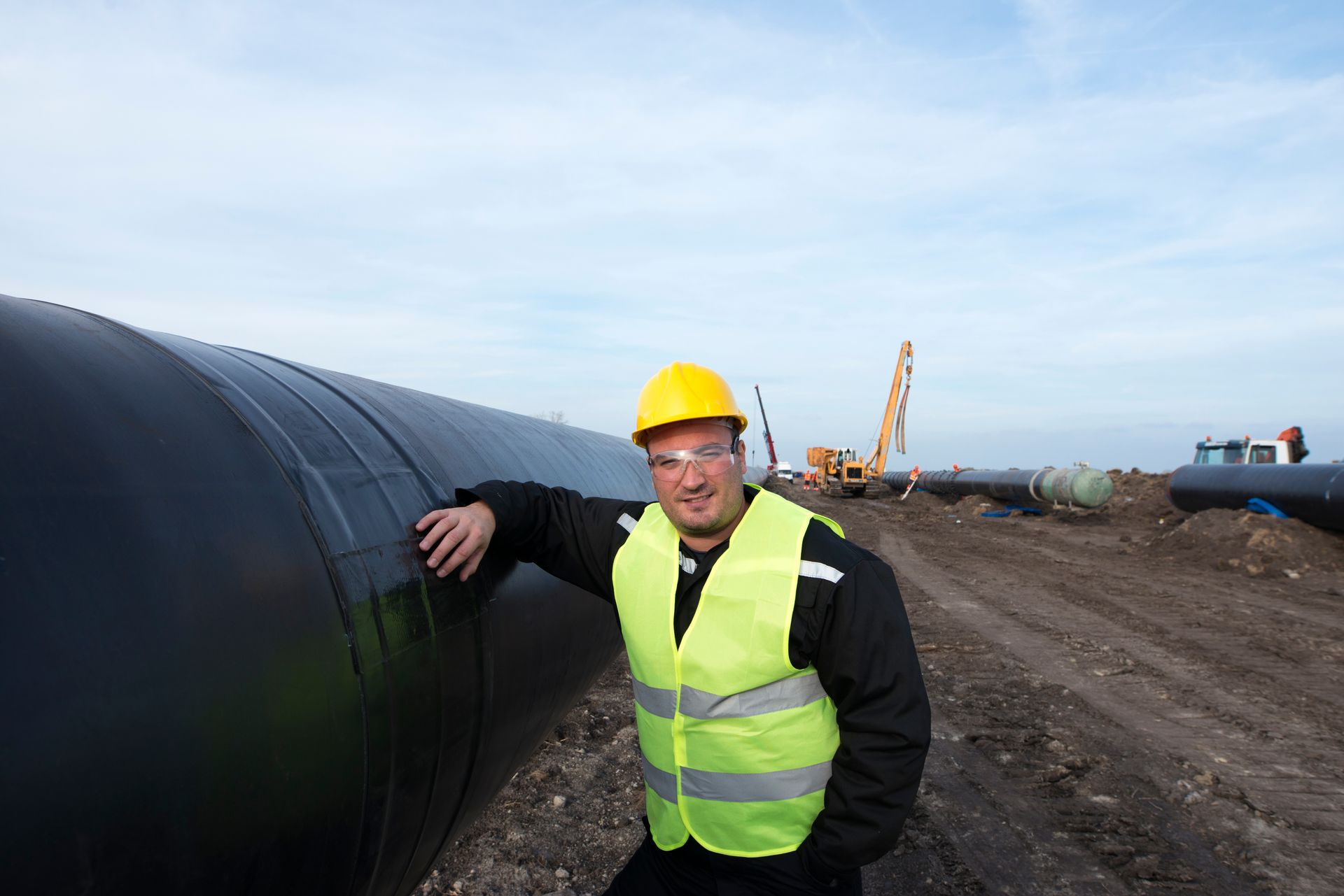Construction worker leaning on large pipe; wearing yellow vest and hard hat. Construction site setting.
