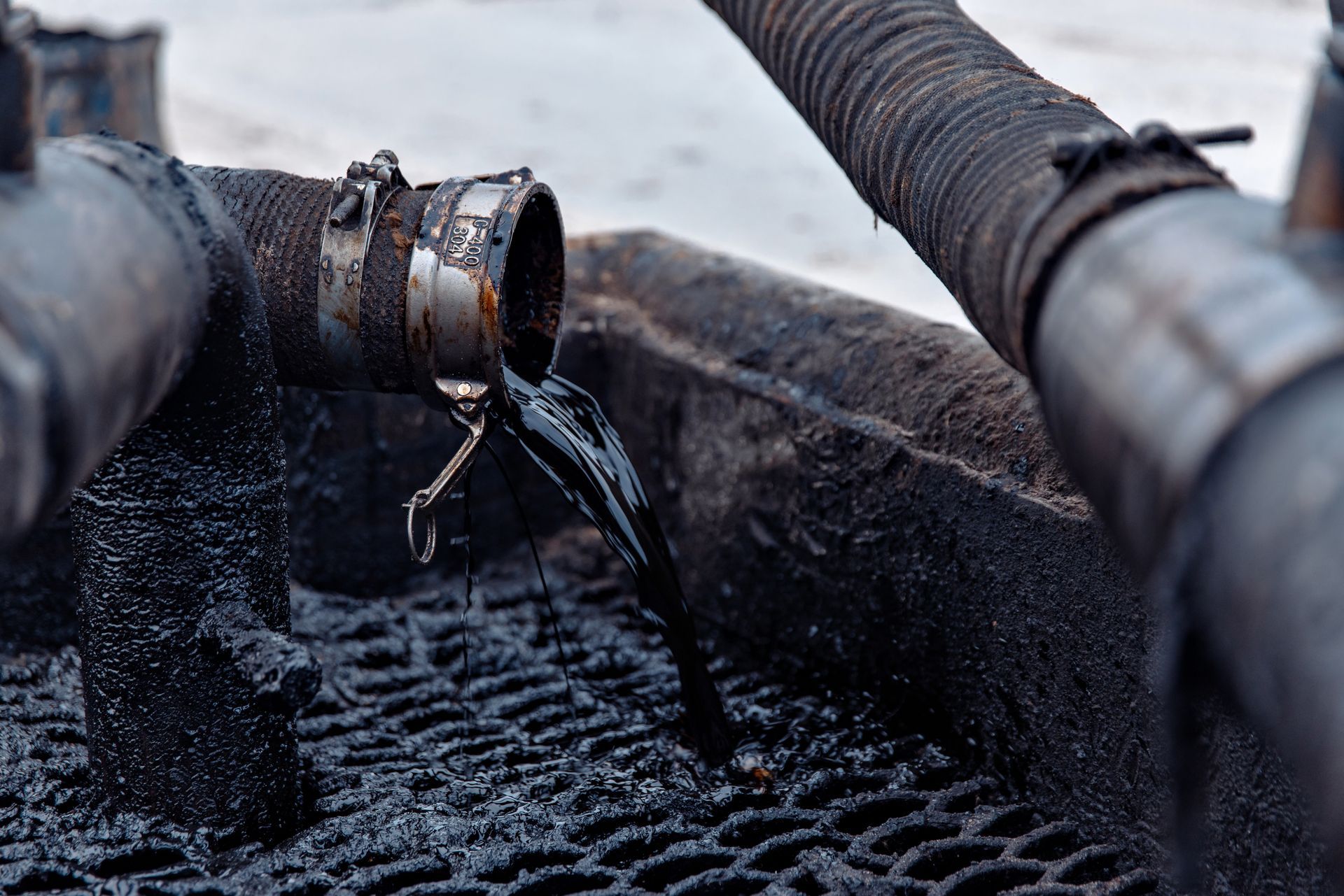 Dark liquid pouring from a pipe, likely oil. Pipe and surrounding area are coated in the same dark substance.