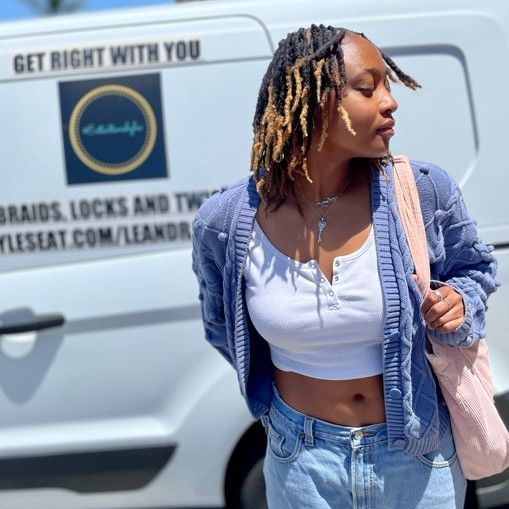 Woman in blue cardigan and jeans, standing near a van with a logo for hair braiding services.