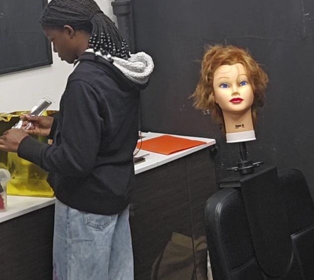 Person in braids prepares supplies near a mannequin head with red hair in a salon.
