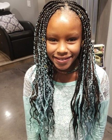 Girl with box braids, some with blue-gray tips, smiles in a salon, wearing a light blue shirt.