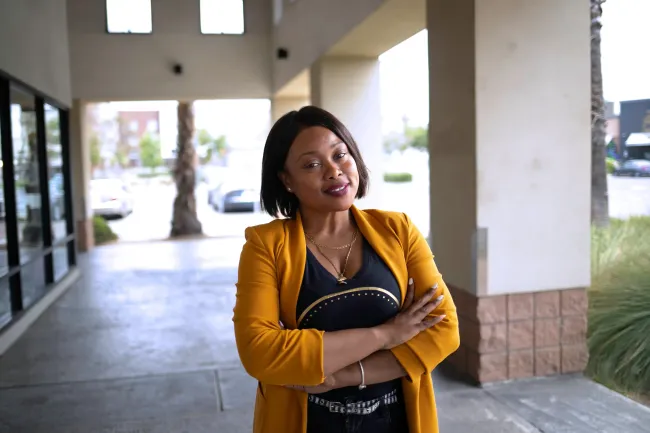 Woman in mustard blazer smiles with arms crossed under a covered walkway.