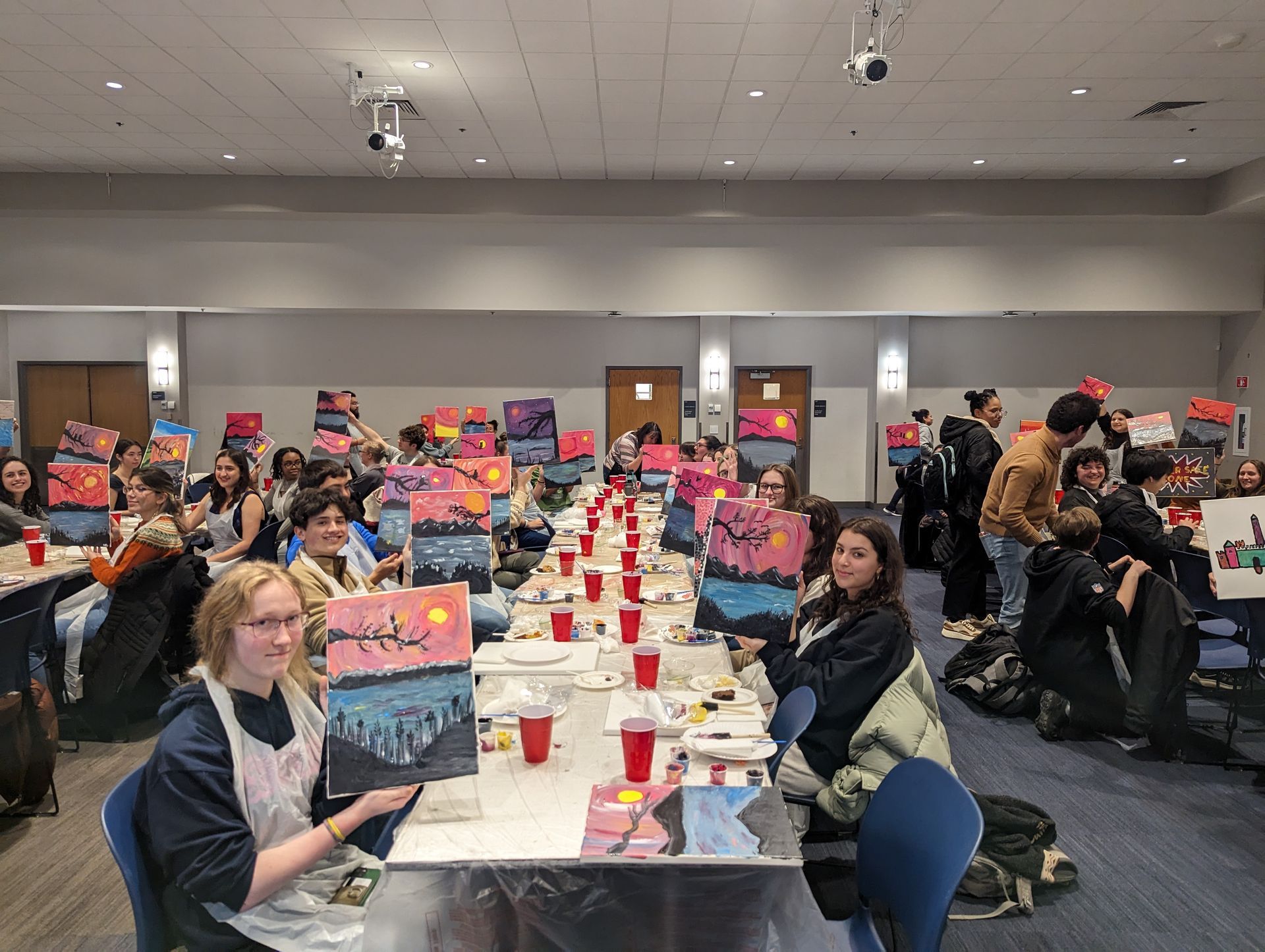 A group of people are sitting at long tables in a room with paintings on them.