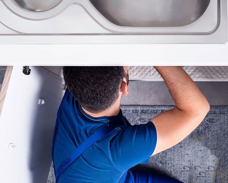 a man in blue overalls fixing a sink in a residential kitchen.