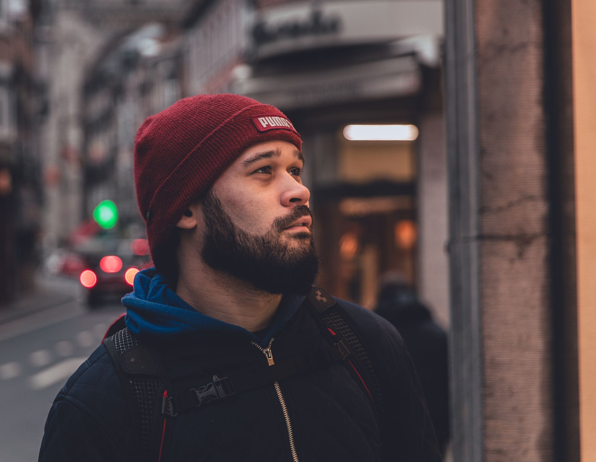 Close-up van een vriend in een opvallende rode beanie, detailrijk tegen de levendige achtergrond van Dinant's regenachtige stadsleven.