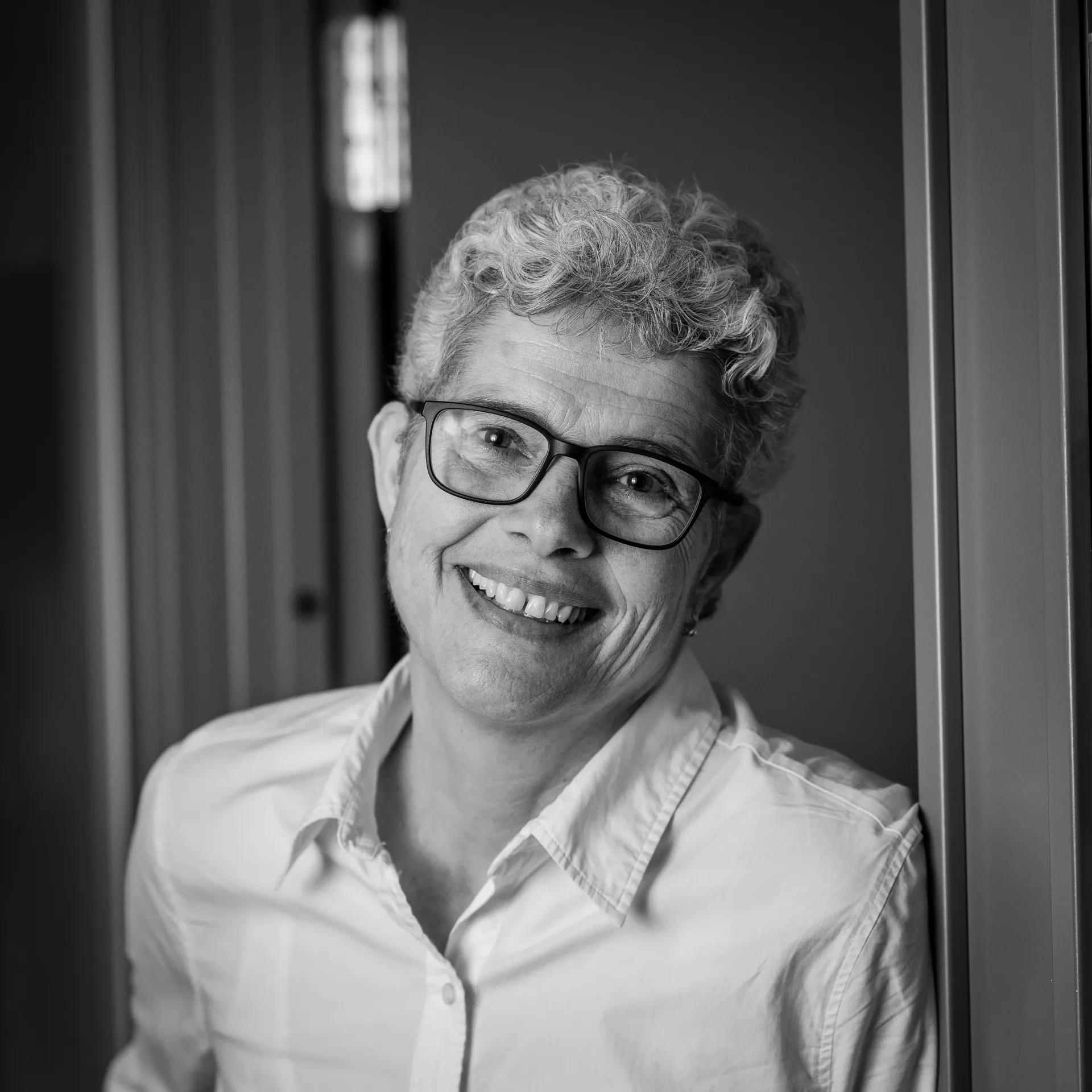 Person med briller, smilende, lent mot en vegg, i et svart-hvitt-bilde.