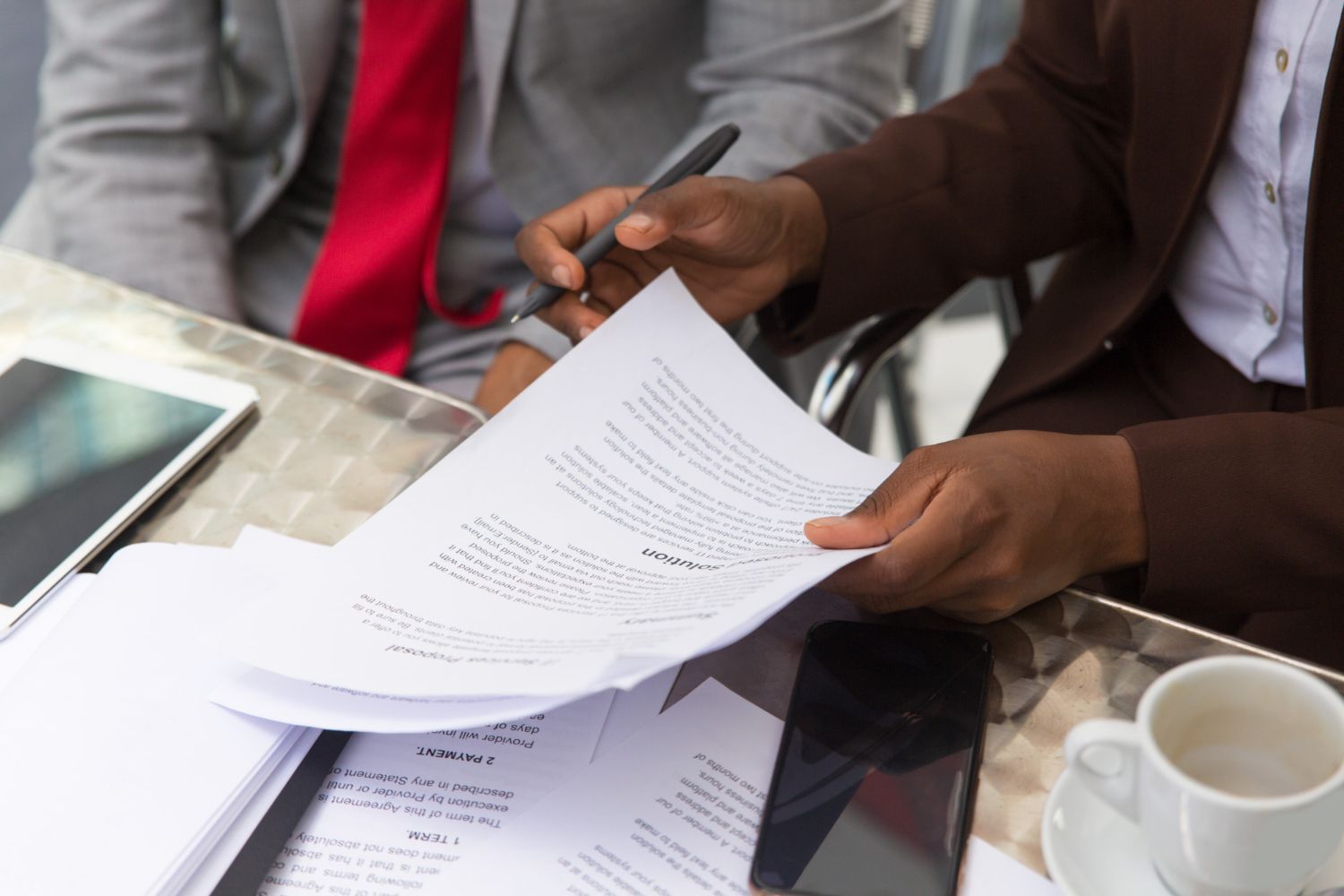 Hands holding documents, one writing with a pen. Papers, tablet, and coffee cup on a table.