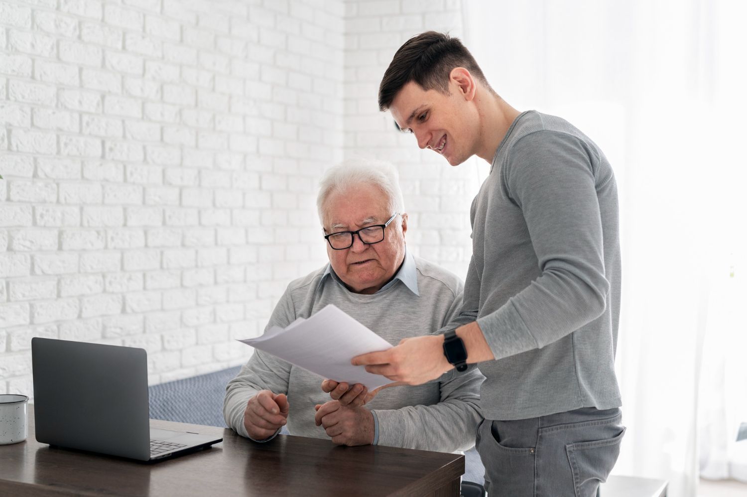 Man assisting older person with paperwork; laptop on table. They both look at the document.