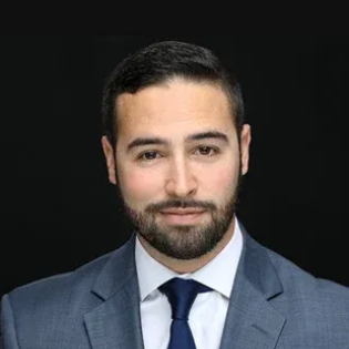 Man in a blue suit and tie, smiling, against a dark background.