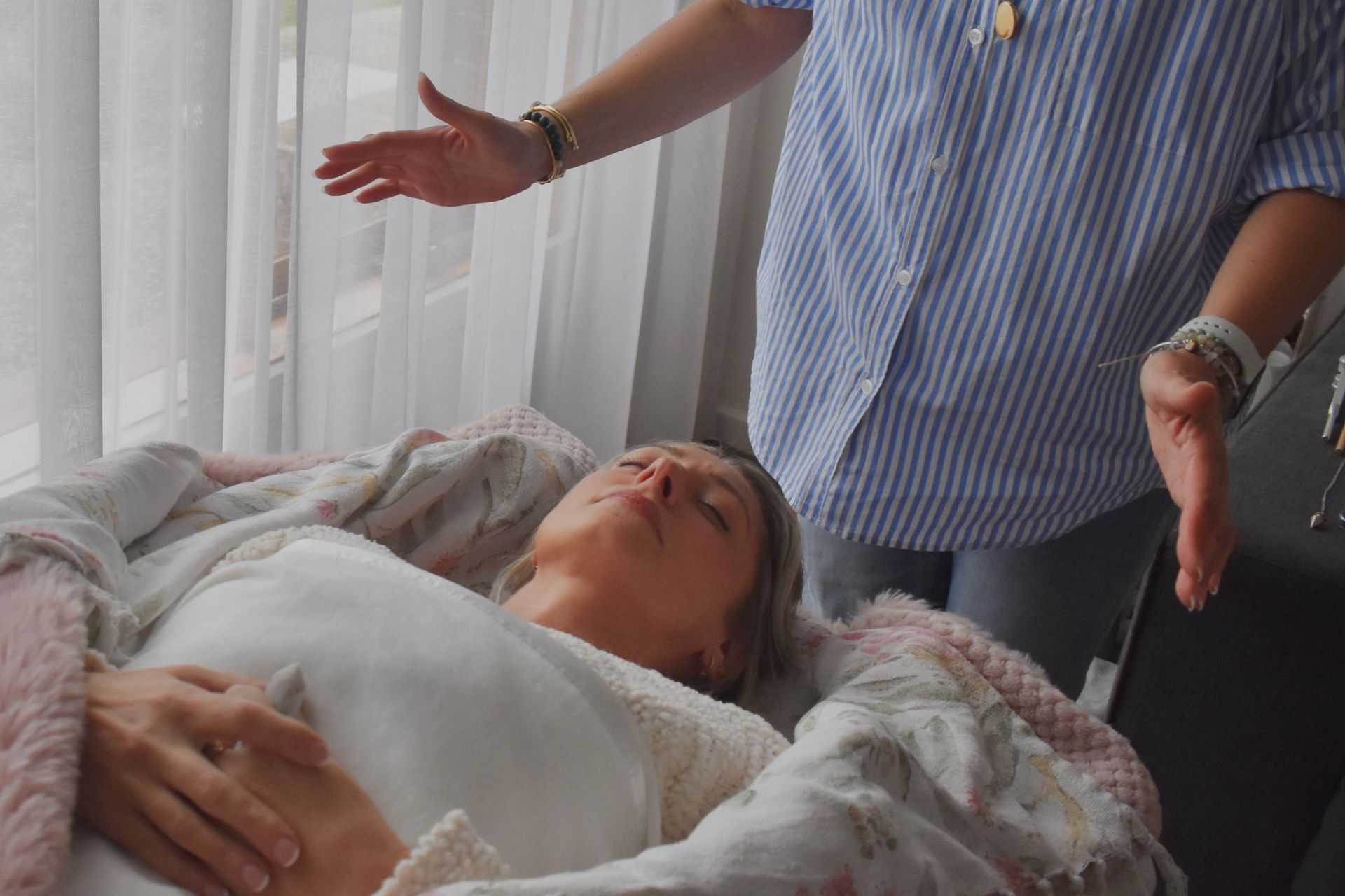 Woman lying down, eyes closed, receiving energy healing. Another person standing with hands outstretched.