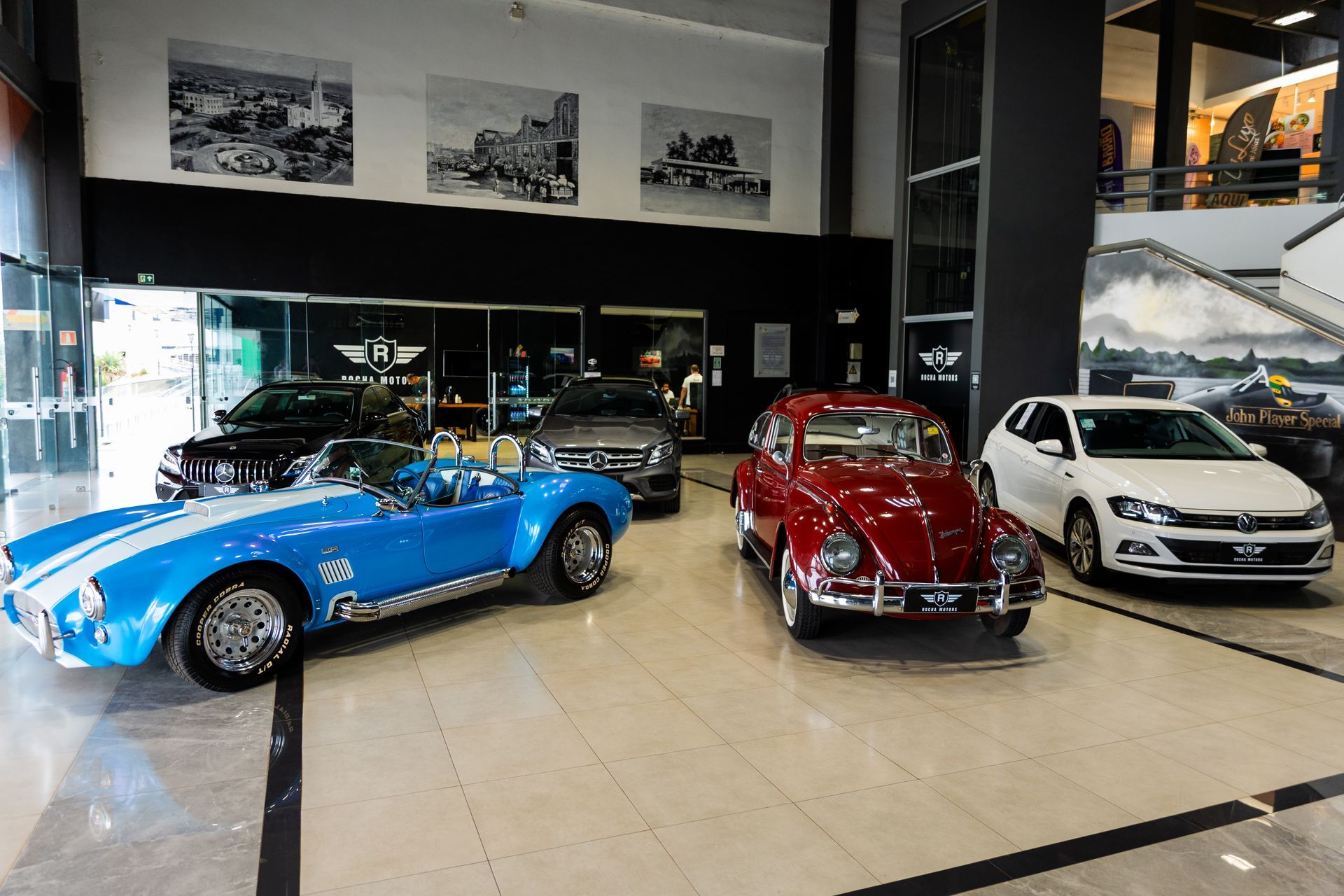 Carros expostos dentro de um showroom: conversível azul, carro clássico vermelho e hatchback branco.