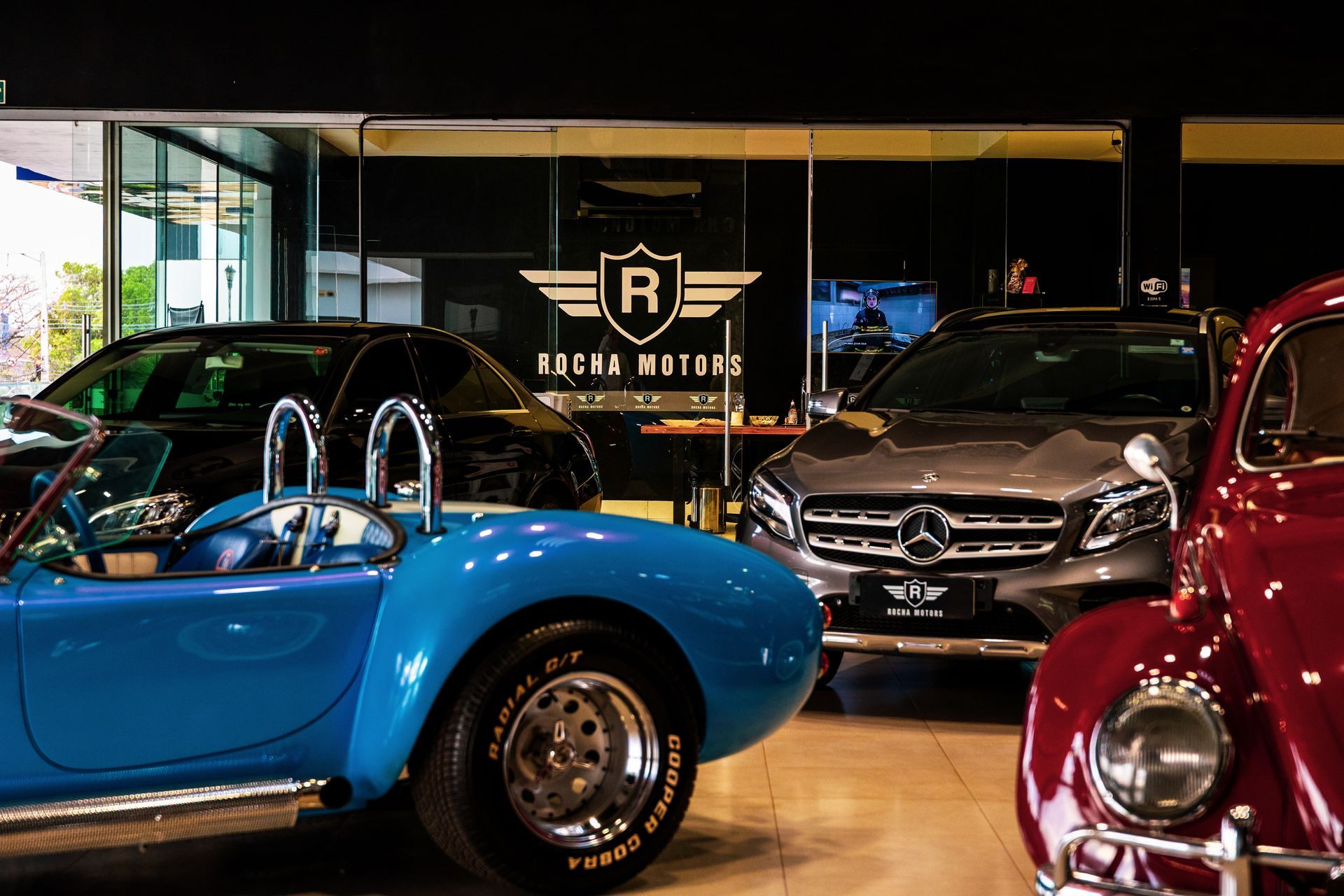 Carros em exposição dentro de um showroom: um conversível azul, um Mercedes cinza e um Volkswagen Beetle