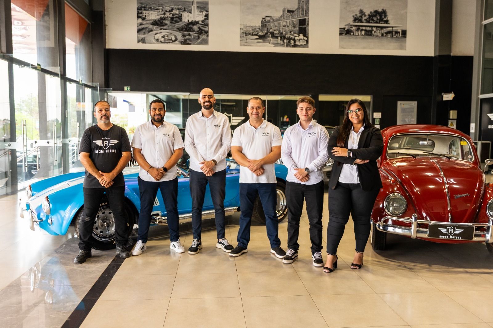 Grupo de sete pessoas posando com carros clássicos em um showroom. Veículos azuis e vermelhos.