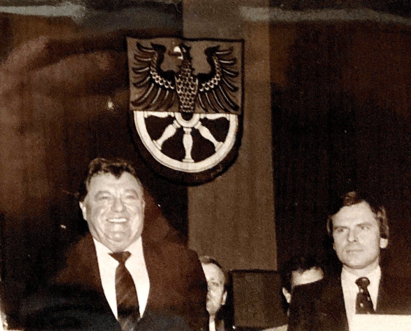 Ein Mann in Anzug und Krawatte steht vor einem Wappen