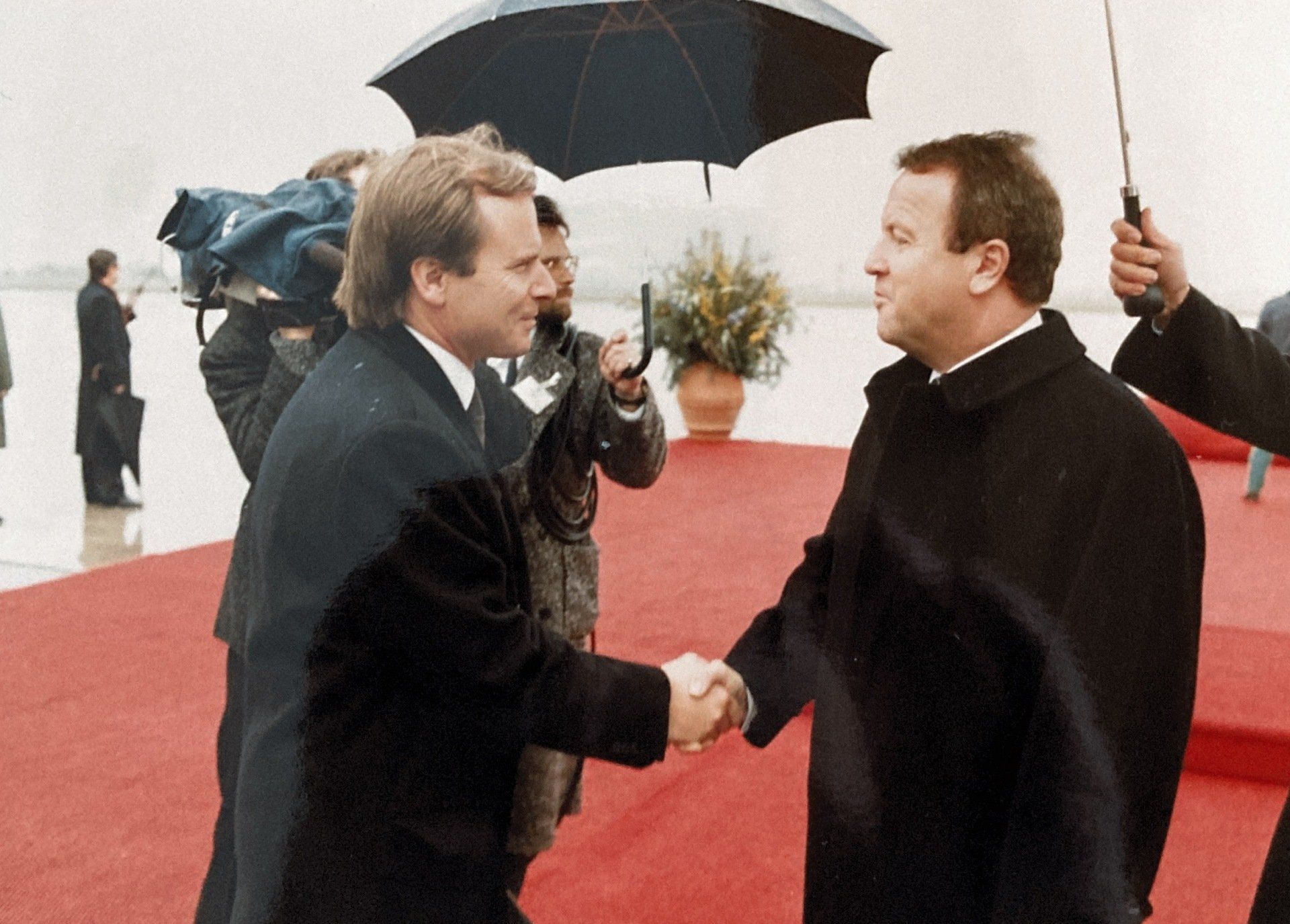 Zwei Männer schütteln sich auf einem roten Teppich im Regen die Hände