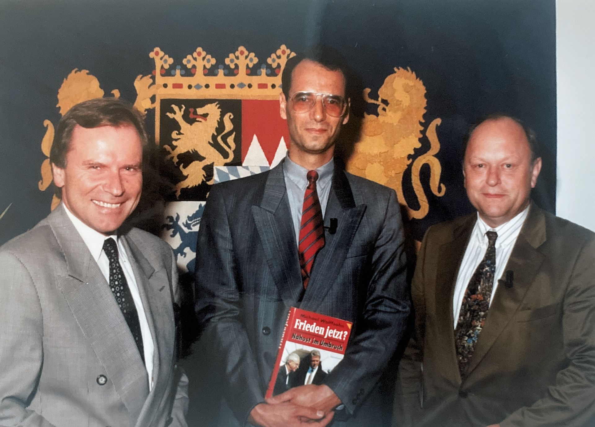 Drei Männer in Anzug und Krawatte posieren für ein Foto vor einem Wappen