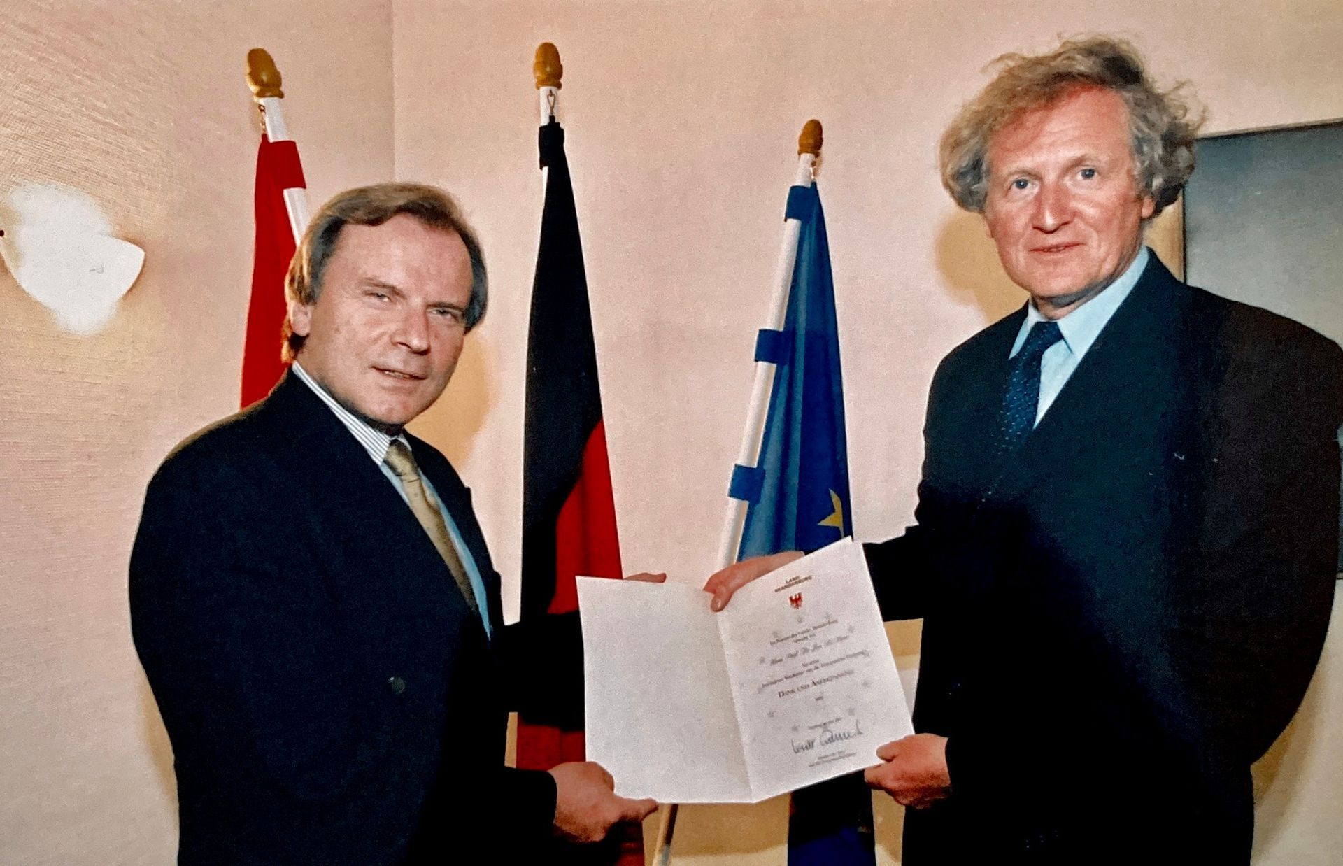 Zwei Männer in Anzügen halten ein Stück Papier vor Flaggen