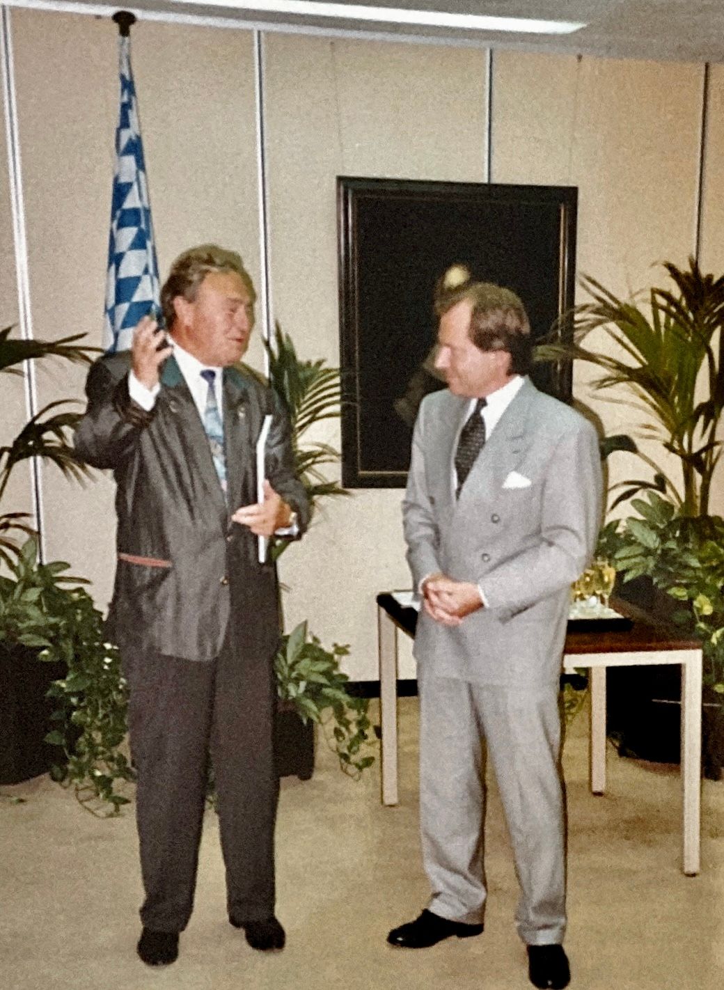 Zwei Männer in Anzügen stehen nebeneinander vor einer Flagge