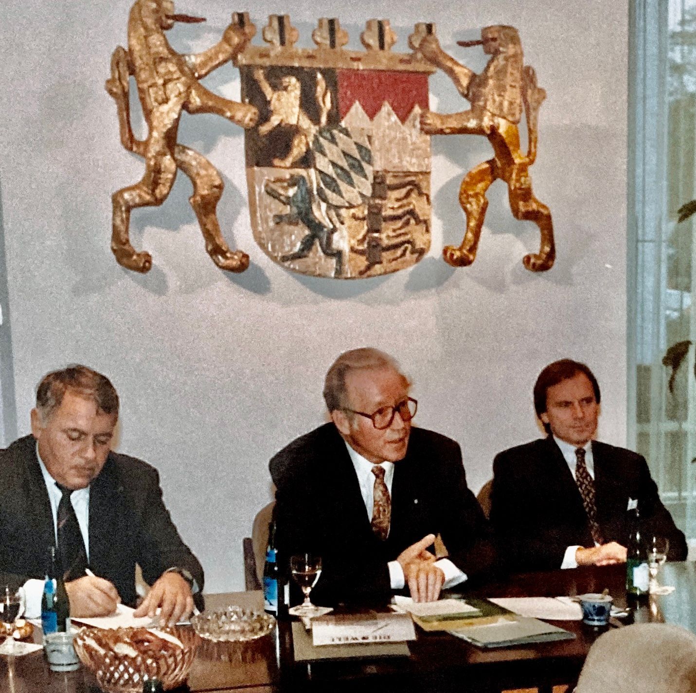 Drei Männer sitzen an einem Tisch vor einem Wappen