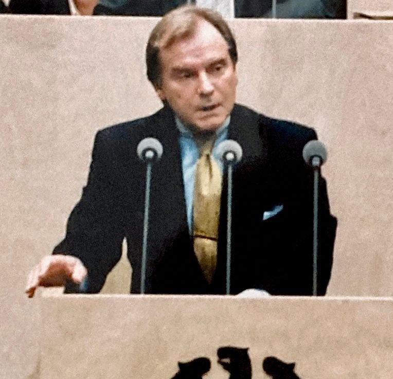 Ein Mann in Anzug und Krawatte hält eine Rede vor Mikrofonen