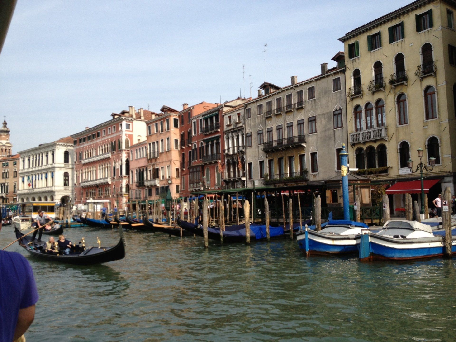 Ein Mann in einem lila Hemd steht vor einem Kanal mit Booten und Gebäuden