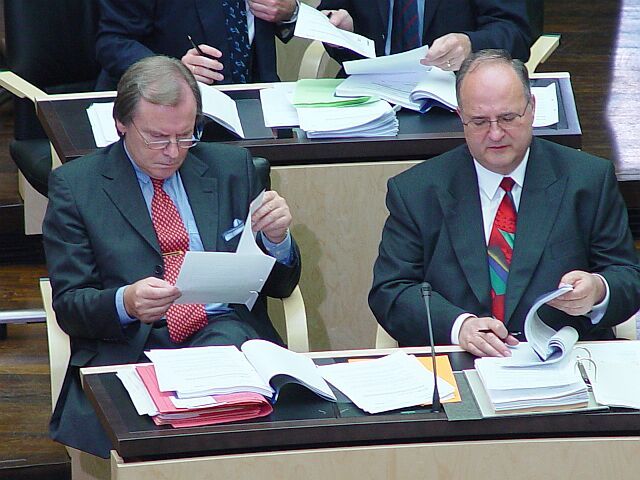 Zwei Männer in Anzug und Krawatte sitzen an einem Tisch und schauen sich Papiere an