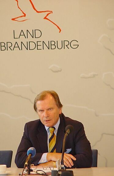 Ein Mann sitzt an einem Tisch mit zwei Mikrofonen vor einer Wand mit der Aufschrift „Land Brandenburg“