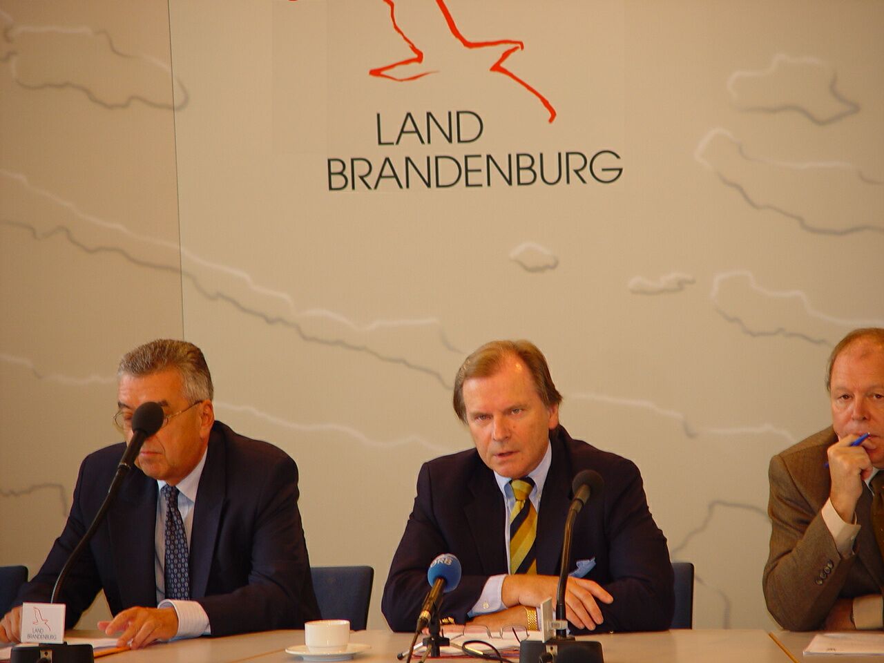 Drei Männer sitzen an einem Tisch mit Mikrofonen vor einer Wand mit der Aufschrift „Land Brandenburg“