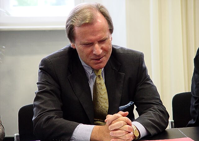 Ein Mann in Anzug und Krawatte sitzt mit gefalteten Händen an einem Tisch.