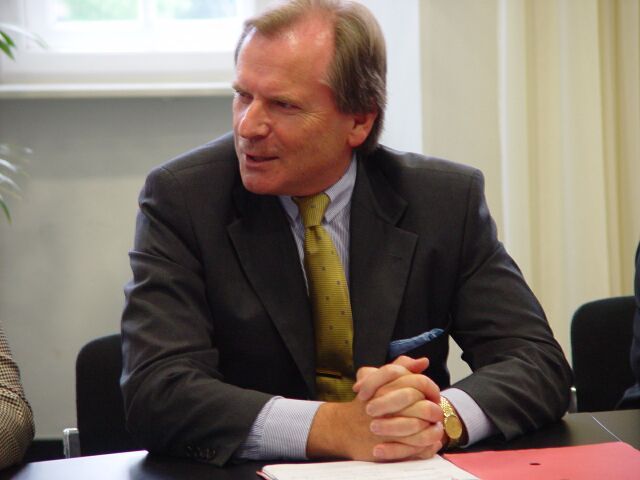 Ein Mann in Anzug und Krawatte sitzt mit gefalteten Händen an einem Tisch