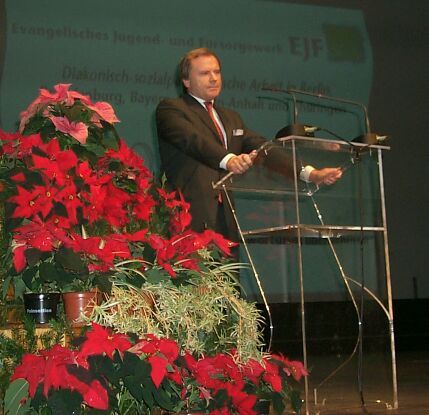 Ein Mann in Anzug und Krawatte steht an einem Podium, umgeben von roten Blumen