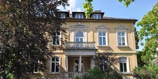 Ein großes Haus mit Balkon ist an einem sonnigen Tag von Bäumen umgeben.