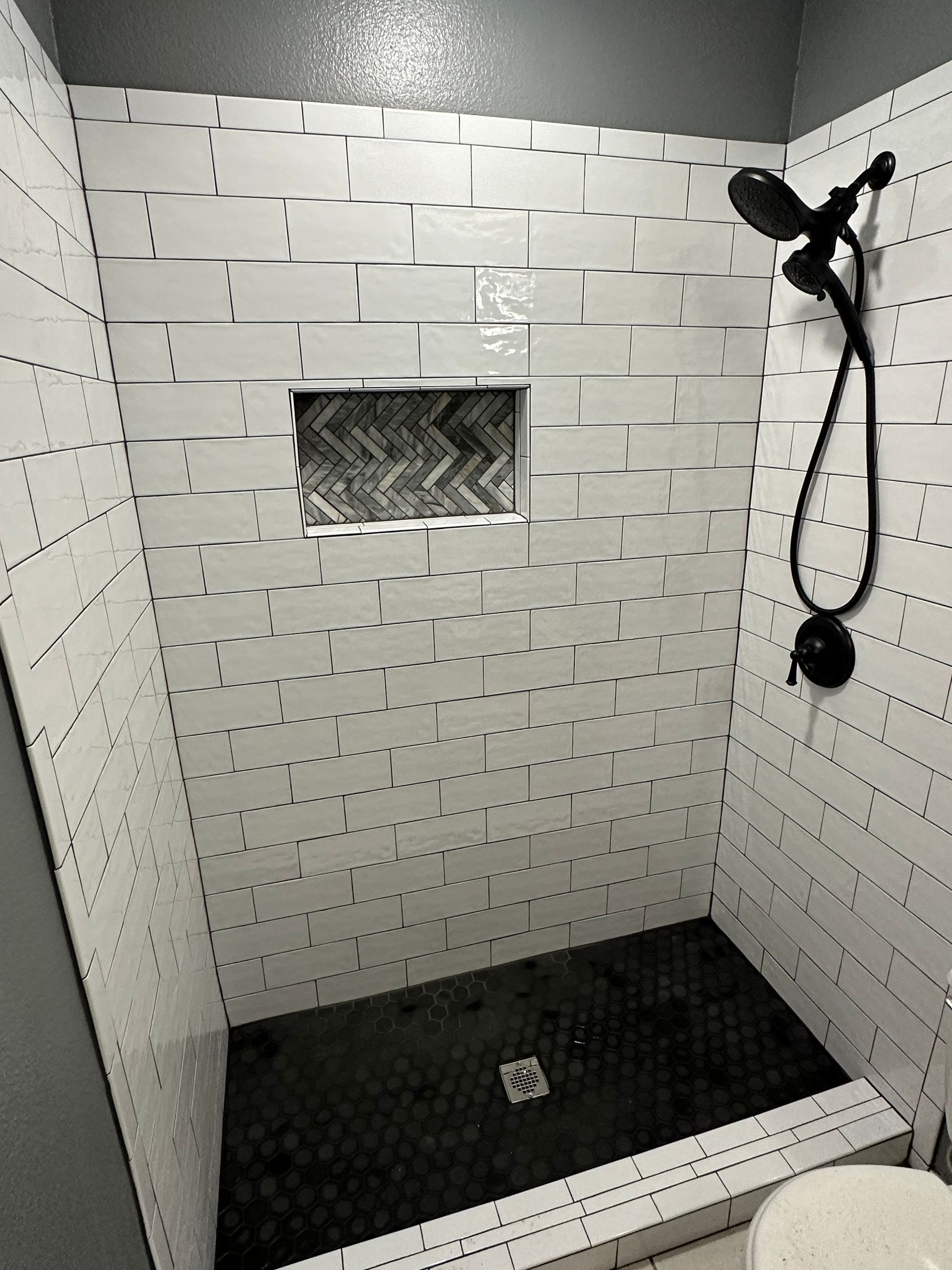 White tiled shower with dark floor and black showerhead, featuring an inset shelf with textured tiles.