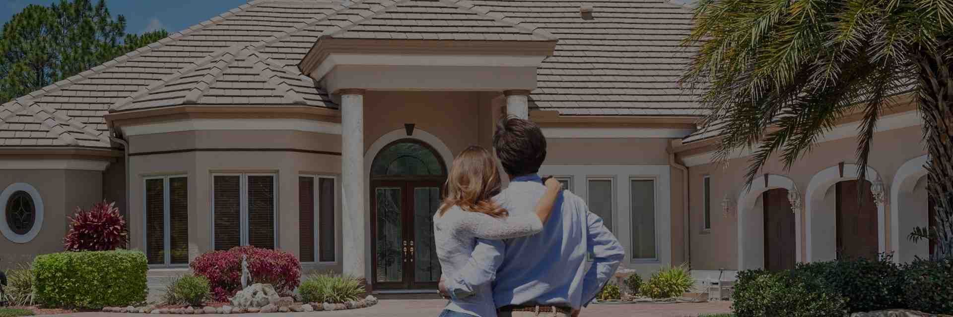 Couple embracing in front of a large house with arched doorway and shrubs in the yard