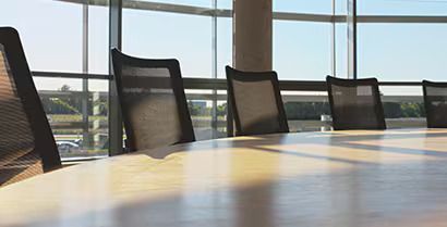 Empty conference room with black chairs around a sunlit wooden table and large windows overlooking greenery