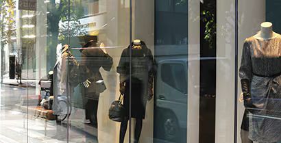 Mannequins in a clothing store window display dark coats and a black dress, with street reflections.