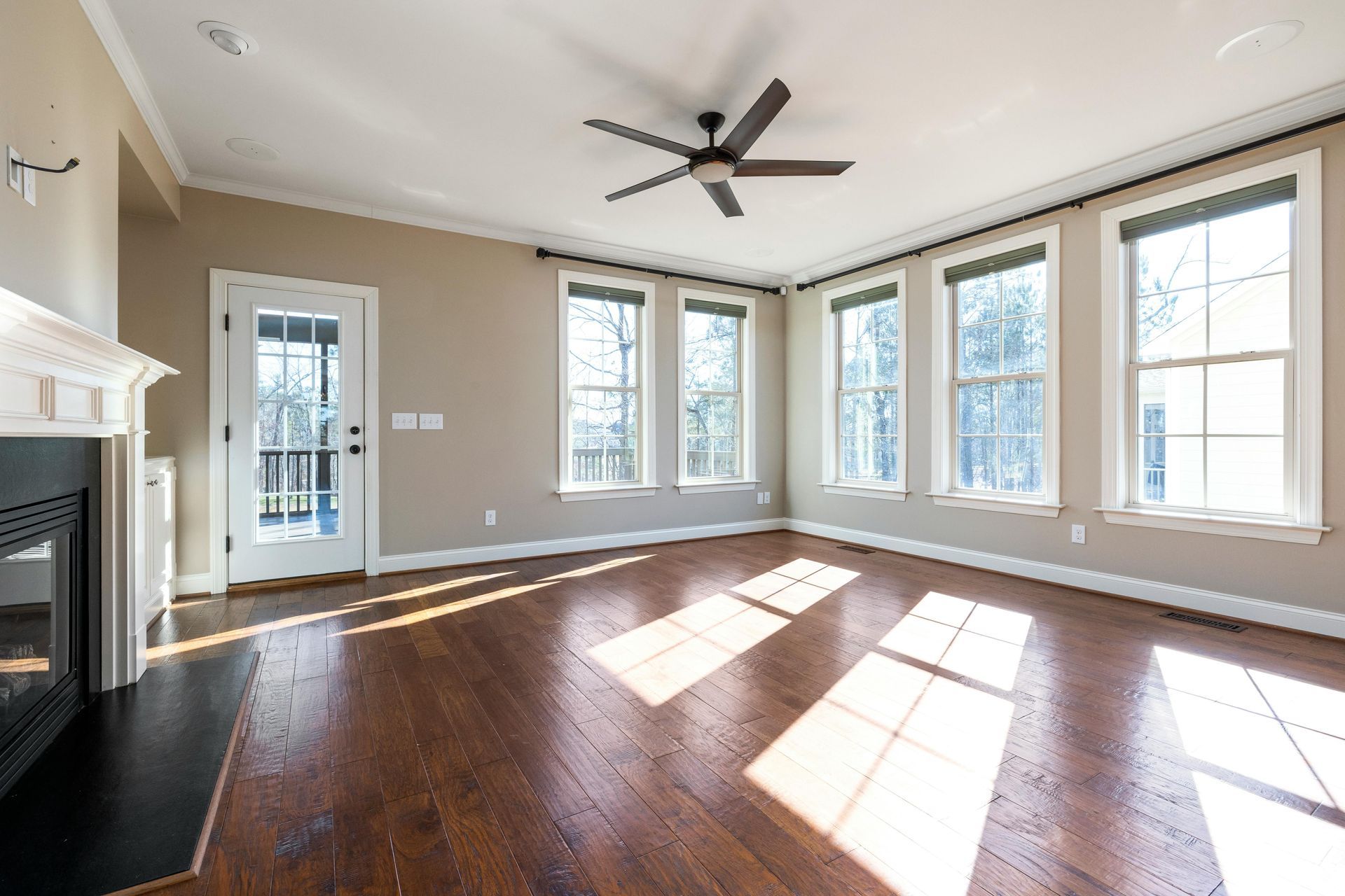 Bright empty living room with dark hardwood floors, large windows, ceiling fan, and fireplace
