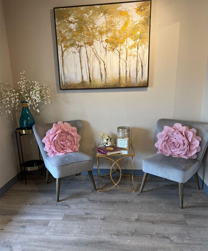 Waiting area with two gray chairs, pink flower pillows, gold side tables, and floral art.