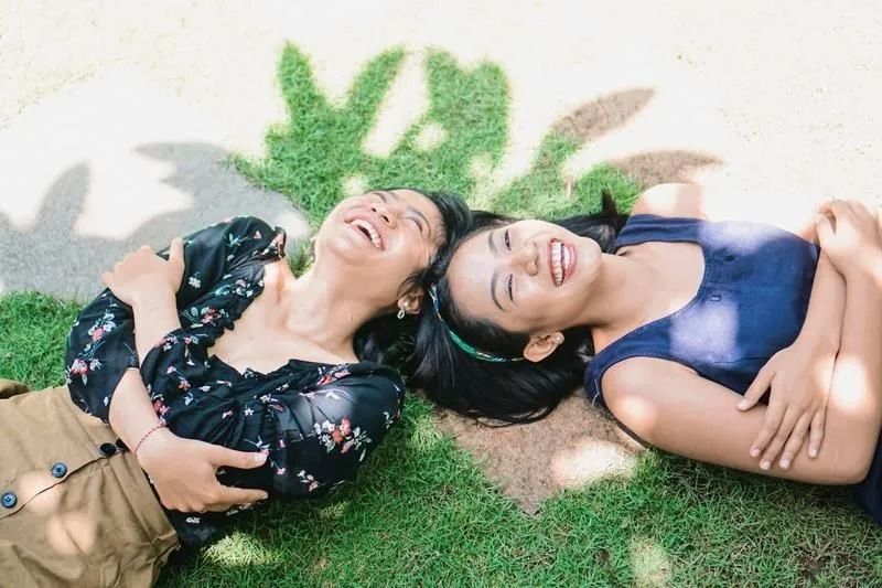 Two laughing people lying on grass, with shadow of greenery above them.