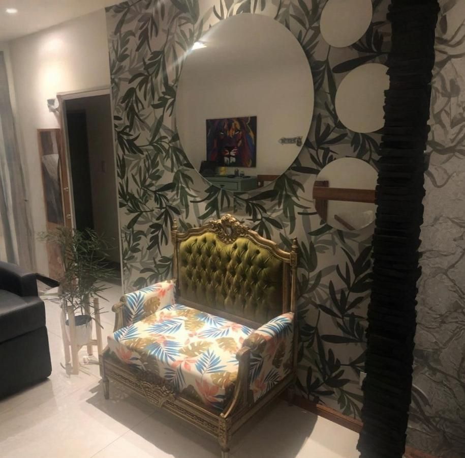 Silla ornamentada en una habitación con papel pintado estampado de hojas, espejo redondo y una columna negra texturizada.