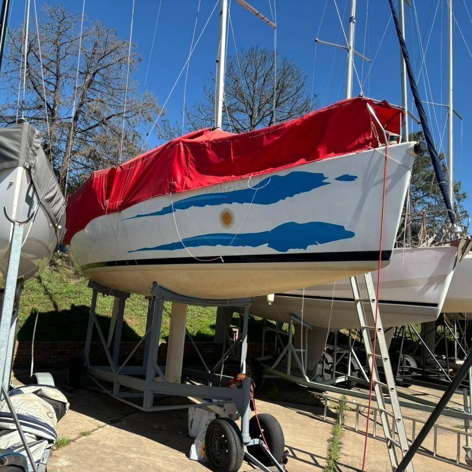 Velero blanco sobre remolque con diseños de olas azules y lona roja; estacionado al aire libre.